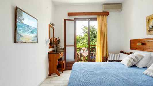 Et hotelværelse med en blå seng, et åbent vindue mod en altan og grønne træer udenfor. (AI-genereret tekst)