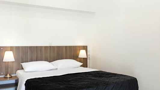 Et soveværelse med en dobbeltseng, hvidt sengetøj og et mørkt sengetæppe. To sengelamper står på sengeborde ved siden af sengen. (AI-genereret tekst)