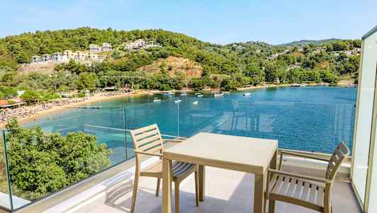 Udsigt fra en terrasse med stole og bord mod en bugt med klart blåt vand, omgivet af grønne bakker og bygninger. (AI-genereret tekst)