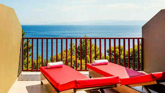 To røde solsenge med hvide håndklæder på en altan med udsigt over havet og vegetation. (AI-genereret tekst)