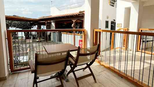 En terrasse med to træstole og et bord. Udsigt over en bygning og et tag. I baggrunden ses en del af havet. (AI-genereret tekst)