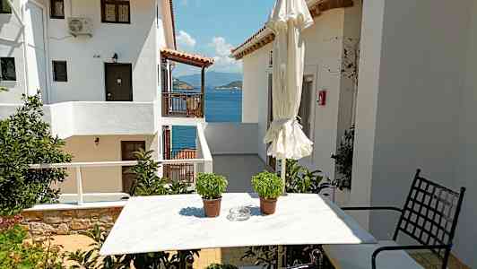 En hvid husbygning med udsigt over havet, et havebord med to krukker og en sammenslået parasol. (AI-genereret tekst)