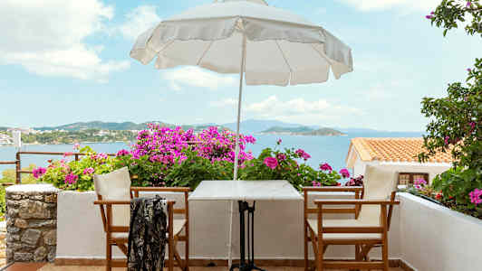 En terrasse med bord, stole, en parasol og blomster, med havudsigt. (AI-genereret tekst)
