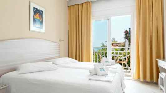 Et lyst hotelværelse med to enkeltsenge, store vinduer med gulddraperier og adgang til en altan. (AI-genereret tekst)