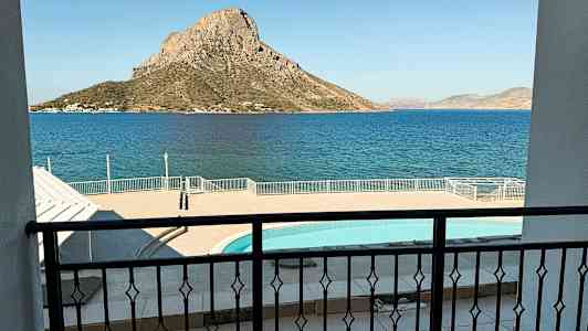 Billedet viser udsigten fra en altan over en pool og havet, med en bjergagtig ø i baggrunden. (AI-genereret tekst)