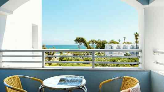 Udsigt fra en altan med havet i baggrunden. En gul stol og et bord er på altanen. (AI-genereret tekst)