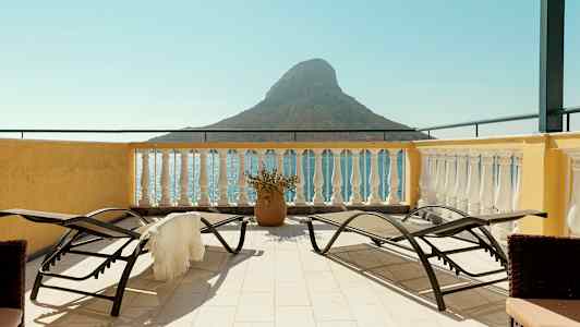 Terrasse med liggestole og udsigt over havet og en lille bjergformation. (AI-genereret tekst)