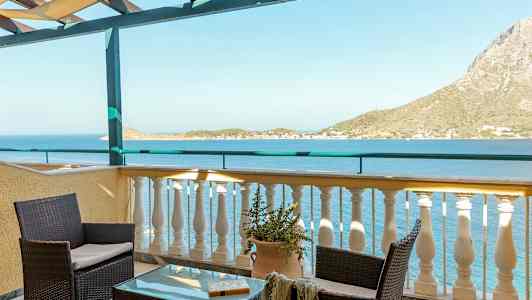 Udsigt fra en terrasse med bord og stole, der vender mod havet og en bjergrig kyst. (AI-genereret tekst)