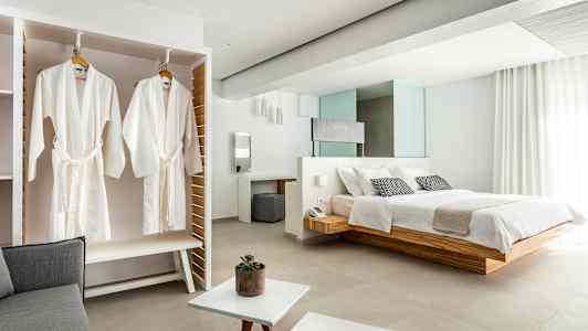 Billede af et elegant hotelværelse med en stor seng, hvide badekåber hængende i et skab og lyse moderne møbler. (AI-genereret tekst)