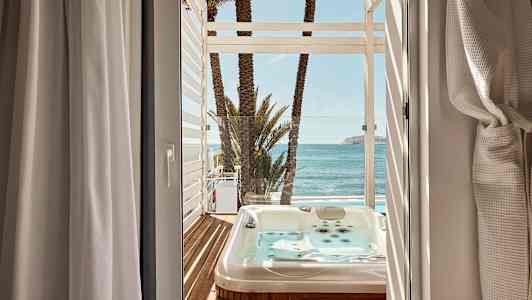 Udsigt fra et værelse mod en terrasse med boblebad, palmer og hav. (AI-genereret tekst)