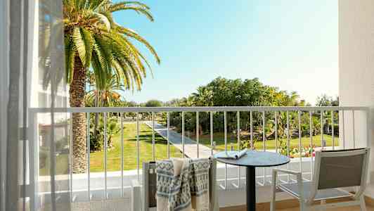 Udsigt fra en altan med et bord, stole og et håndklæde, mod en have med palmer og gangsti. (AI-genereret tekst)