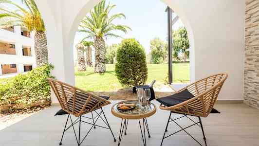 To flettede stole og et bord med glas og en tallerken på en terrasse, med udsigt over en grøn have med palmer. (AI-genereret tekst)