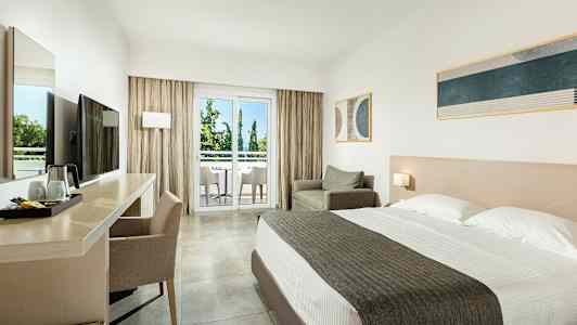 Et hotelværelse med dobbeltseng, skrivebord og stol, lænestol samt en altan med udsigt til træer. (AI-genereret tekst)