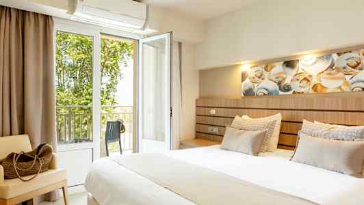 Et hotelværelse med en stor seng, siddemøbel og åben altandør med udsigt til grøn beplantning. (AI-genereret tekst)