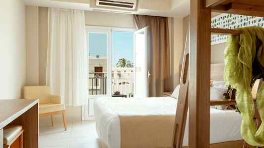 Et hotelværelse med en seng, et skrivebord og en stol, samt et åbent glasvindue ud mod en altan. (AI-genereret tekst)