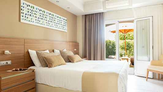 Et lyst hotelværelse med en stor seng, vægkunst og altandøre med udsigt til en have med en orange parasol. (AI-genereret tekst)