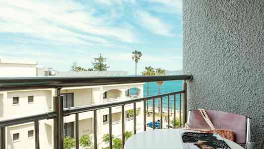 Udsigt fra en balkon mod stranden med havet og palmer i baggrunden. (AI-genereret tekst)