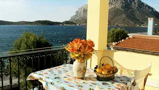 En udsigt fra en altan mod havet, med et bord dekoreret med en vase med orange blomster og en kurv med citroner. (AI-genereret tekst)