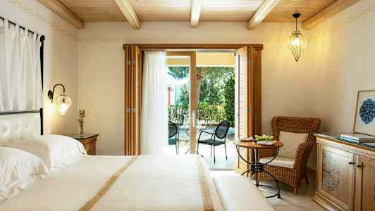 Et soveværelse med dobbeltseng, lyse møbler og glasdøre, der fører til en møbleret udendørs terrasse. (AI-genereret tekst)