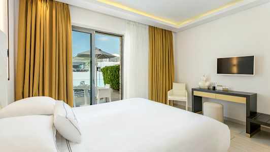 Hotelværelse med hvidt interiør, en dobbeltseng, skrivebord og udgang til terrasse. (AI-genereret tekst)