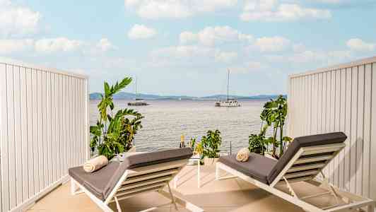 To solsenge med grå puder og håndklæder placeret på en terrasse med udsigt over havet og sejlbaåde. (AI-genereret tekst)
