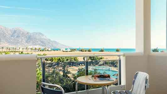 Udsigt fra en balkon med bord og stole mod en pool, landskab med bjerge og havet i baggrunden. (AI-genereret tekst)