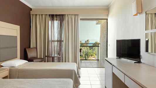 Hotelværelse med to senge, tv, bord og stole samt en balkon med udsigt over grønne områder og hav. (AI-genereret tekst)