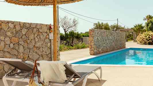 To solsenge med et håndklæde og en stråtaske ved siden af en pool. Der er også en stenmur og palmer i baggrunden. (AI-genereret tekst)