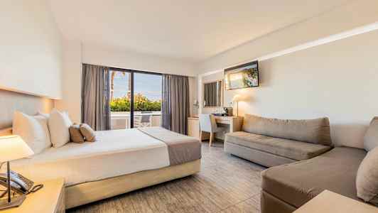 Et hotelværelse med en seng, sofaer, skrivebord med spejl og en siddegruppe nær et glasvindue. (AI-genereret tekst)