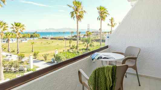 En balkon med bord, stole og udsigt over en grøn have med palmer samt havet i baggrunden. (AI-genereret tekst)