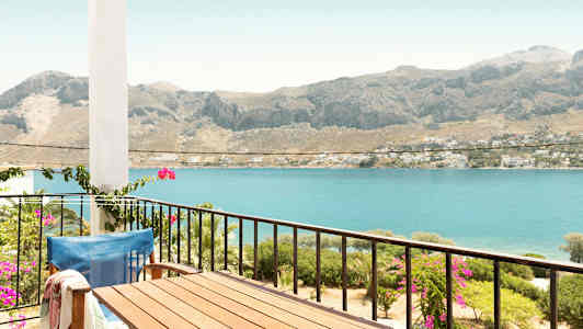 En balkon med bord og stol med udsigt over havet og bjerge i baggrunden. (AI-genereret tekst)