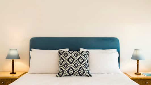 En seng med en blå sengegavl, hvide puder og en dekorativ pude i midten. To natborde med lamper på hver side. (AI-genereret tekst)