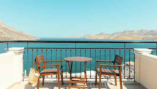 To træstole og et bord på en terrasse med udsigt til havet og bjerge i det fjerne. (AI-genereret tekst)