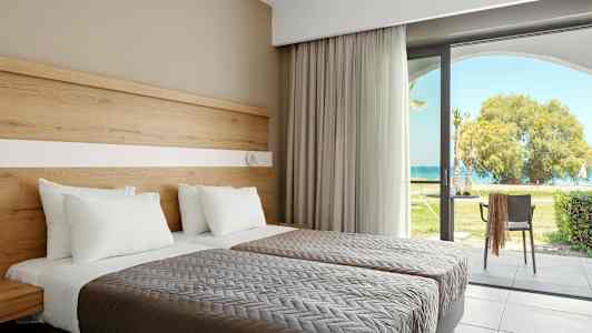 To senge med hvide puder og gråt sengetæppe, ved siden af en glasdør, der fører til en have med udsigt over havet. (AI-genereret tekst)