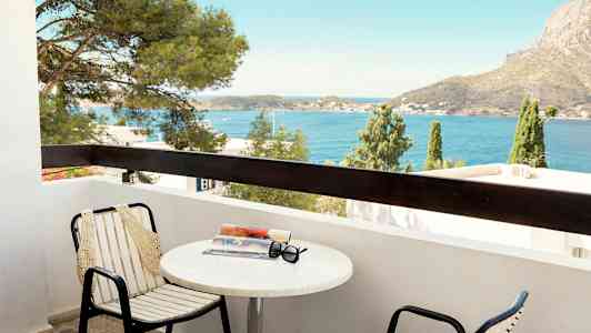 En balkon med stole og bord, hvorpå der ligger en bog og solbriller. Udsigt over havet og en bjergagtig baggrund. (AI-genereret tekst)