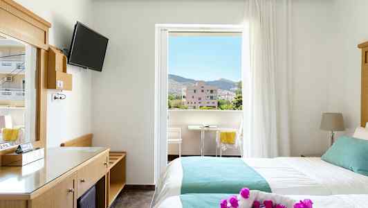 Hotelværelse med to senge og balkon. Udsigt over omkringliggende bygninger og bjerge. (AI-genereret tekst)