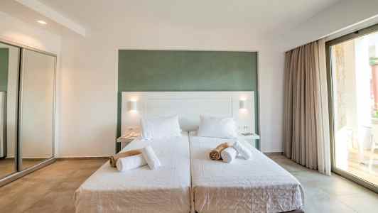 To enkeltsenge med hvidt sengetøj og sammenrullede håndklæder, i et lyst rum med stor glasdør. (AI-genereret tekst)