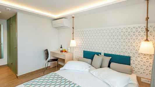 Billedet viser et moderne hotelværelse med en dobbeltseng, hvide vægge og en dekorativ panel bag sengen. Et skrivebord med stol og frugt kan ses i rummet. (AI-genereret tekst)