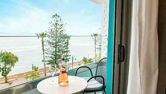 Udsigt fra altan med bord, stole og en flaske rosévin ud mod hav og træer. (AI-genereret tekst)