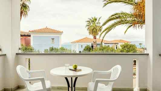 To hvide plaststole og et bord på en altan med udsigt over bygningen med røde tage og palmer. (AI-genereret tekst)