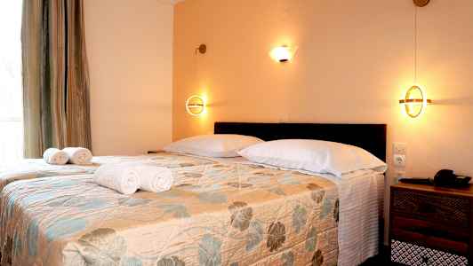 Et soveværelse med to enkeltsenge dækket med et blomstret sengetæppe, hvide puder og rullede håndklæder, samt væglamper. (AI-genereret tekst)