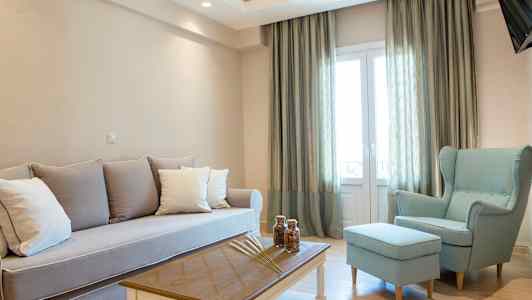 En stue med lyse møbler, herunder en sofa, en lænestol og et sofabord, ved siden af store vinduer med gardiner. (AI-genereret tekst)