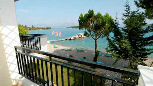 Udsigt fra en altan mod en strand med liggestole ved en klarblå havbugt. Træer og skyer i baggrunden. (AI-genereret tekst)