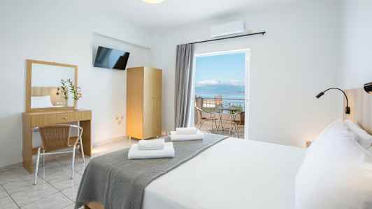 Et lyst hotelværelse med en dobbeltseng, skrivebord, spejl og en altan med udsigt over havet. (AI-genereret tekst)