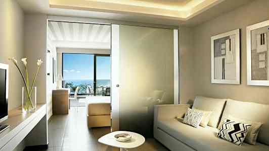 Lyst hotelværelse med sofa, lavt bord og en glasvæg mod soveværelsesdelen med dobbeltseng. Balkon med havudsigt ses. (AI-genereret tekst)