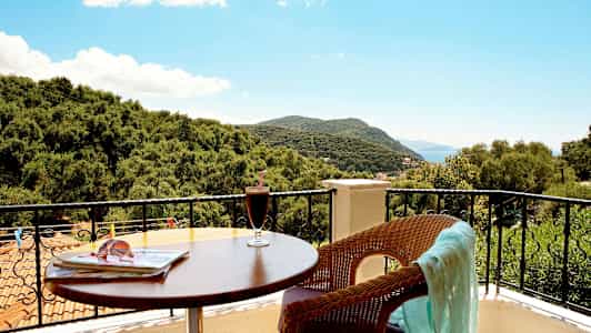 Udsigt fra en balkon med bord, drik og en stol med et klæde samt træer og bjerge i baggrunden. (AI-genereret tekst)