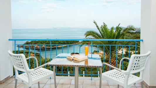 Udsigt fra en altan mod havet, med et bord og to stole i forgrunden. (AI-genereret tekst)