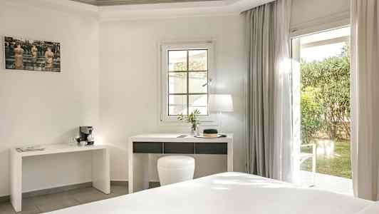 Et lyst hotelværelse med et skrivebord og en vinduesdør ud til en have. (AI-genereret tekst)