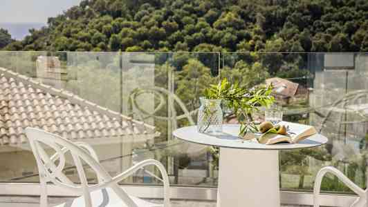 Et bord med glasvase, frugt, bog og glas på en terrasse med udsigt over træer og tage. (AI-genereret tekst)