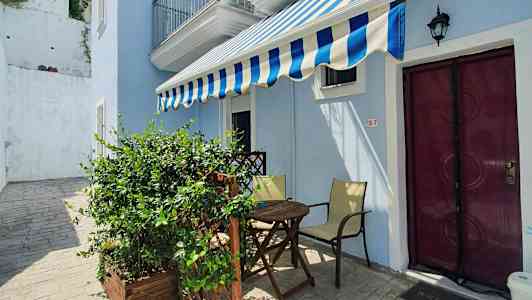 En lille udendørs terrasse med et bord, stole og en markise foran en lyseblå bygning. (AI-genereret tekst)